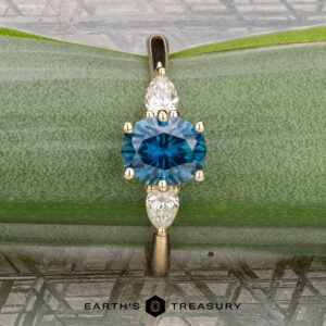 The "Nymph" ring in 14K yellow gold features a 1.27-carat Montana sapphire flanked by two pear-shaped clear stones, beautifully presented on a green leaf background for an enchanting, nature-inspired look.
