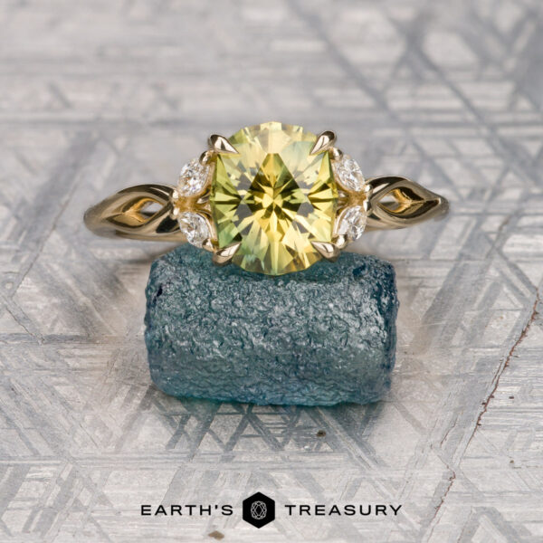 The "Anise" ring in 14K yellow gold with 2.43-carat Australian sapphire The "Anise" ring in 14K yellow gold with a 2.43-carat Australian sapphire features leaf-shaped side accents and is shown on a rough blue-green stone with a patterned gray background. The "Earth's Treasury" logo appears at the bottom.