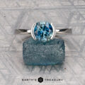 The "Lina" ring in platinum with a 1.83-carat Montana sapphire is displayed on textured blue stone, with the "EARTH'S TREASURY" logo beneath.