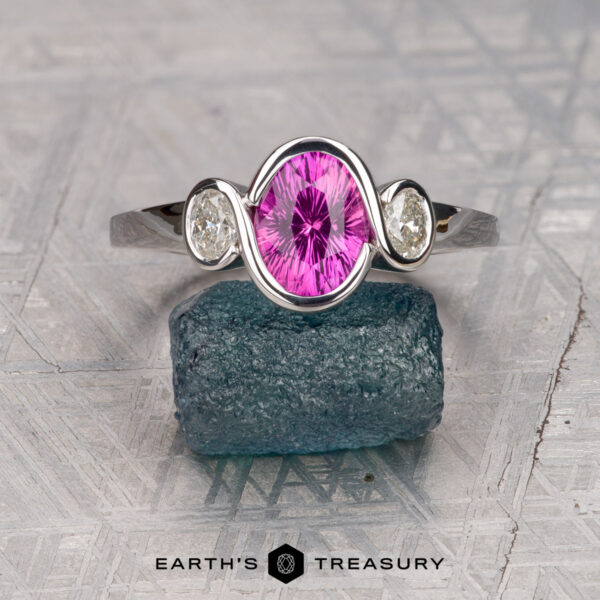 The "Kaelin" ring in 14K white gold with 1.53-carat Ceylon sapphire The "Kaelin" ring in 14K white gold features a 1.53-carat oval Ceylon sapphire flanked by two clear stones, displayed on a rough blue-green crystal with the "Earth's Treasury" logo below.