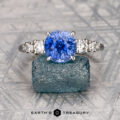 The "Celosia" ring in 18K white gold features a 3.06-carat Ceylon sapphire and diamond accents, shown on a rough gemstone with "Earth's Treasury" branding—a refined, elegant design.