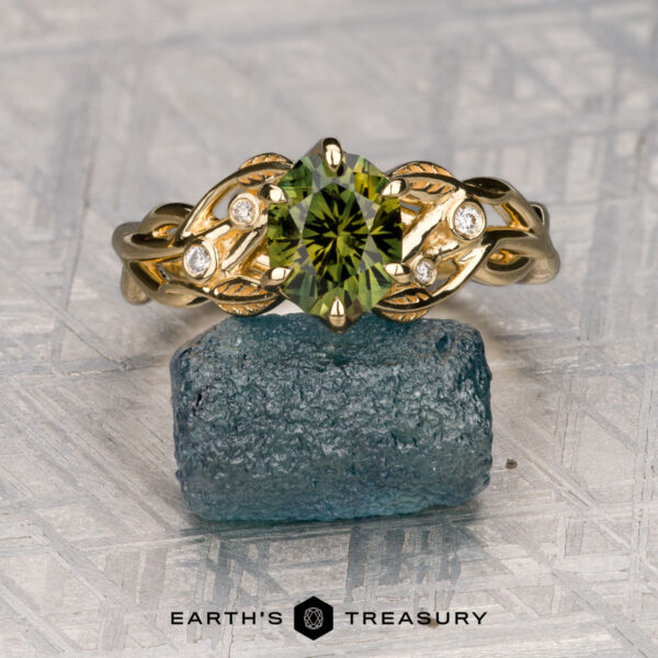 The "Viney" ring in 18k yellow gold with 1.55-carat Australian sapphire The "Viney" ring in 18k yellow gold features a 1.55-carat Australian sapphire, four small diamonds, and leaf motifs on the band; it rests on a rough blue stone with the "Earth's Treasury" logo below.