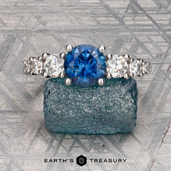 The "Celosia" ring in 14K white gold with 1.30-carat Montana sapphire The "Celosia" ring in 14K white gold with a 1.30-carat Montana sapphire is displayed upright on a blue rough gemstone atop a gray textured surface. The text “Earth’s Treasury” appears below.