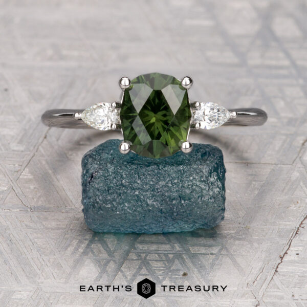 The "Dryad" ring in 18K white gold with 2.21-carat Australian sapphire The "Dryad" ring in 18K white gold, featuring a 2.21-carat Australian sapphire and two clear side stones, is displayed on a rough blue crystal with the "Earth's Treasury" logo at the bottom.