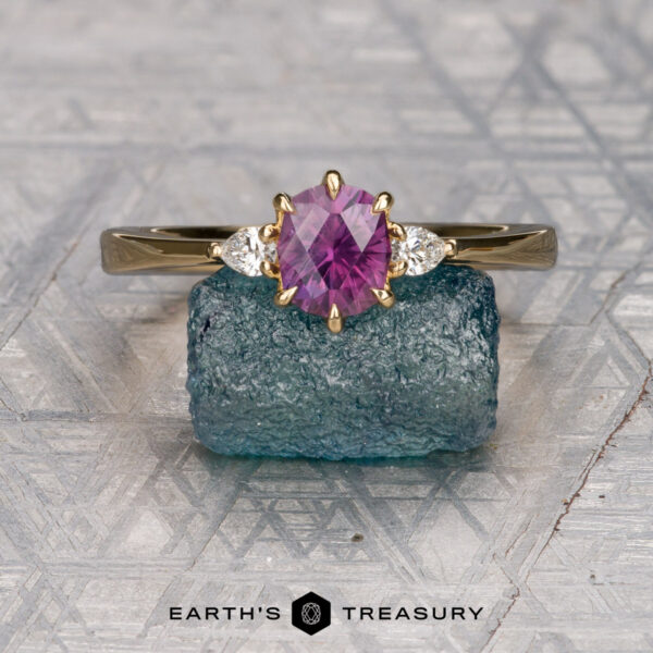 The "Sylph" ring in 18K yellow gold with 0.94-carat Montana sapphire The "Sylph" ring in 18K yellow gold, set with a 0.94-carat Montana sapphire and two white stones, is showcased on a rough blue crystal against a geometric patterned background.