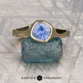 The "Winona" ring in 14K yellow gold features a 1.08-carat Ceylon sapphire, displayed upright on a rough blue stone, echoing solitaire elegance, with the Earth's Treasury logo below.