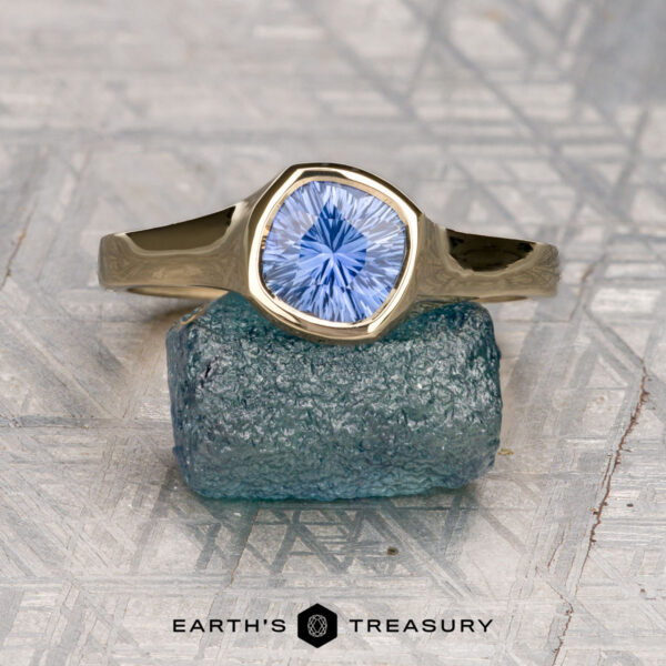 The "Winona" ring in 14K yellow gold with 1.08-carat Ceylon sapphire The "Winona" ring in 14K yellow gold features a 1.08-carat Ceylon sapphire, displayed upright on a rough blue stone, echoing solitaire elegance, with the Earth's Treasury logo below.