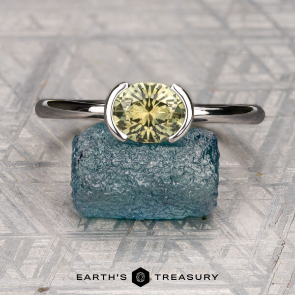 The "Lina" ring in 14K white gold with 1.35-carat Montana sapphire The "Lina" ring in 14K white gold featuring a 1.35-carat Montana sapphire stands upright on a rough blue crystal, set against a geometric patterned background.