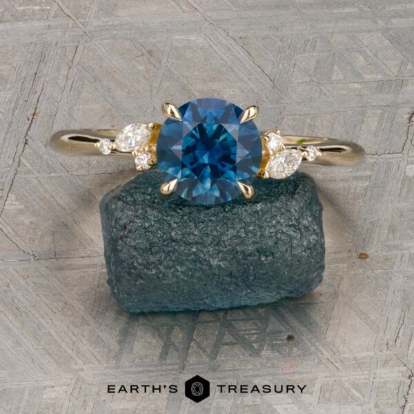The "Iris" ring in 14K yellow gold with 1.60-carat Montana sapphire The "Iris" ring in 14K yellow gold with a 1.60-carat Montana sapphire stands upright against a rough blue stone, with the "Earth's Treasury" logo below on a textured background.