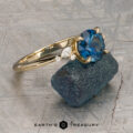 The "Iris" ring in 14K yellow gold with a 1.60-carat Montana sapphire stands upright against a rough blue stone, with the "Earth's Treasury" logo below on a textured background.