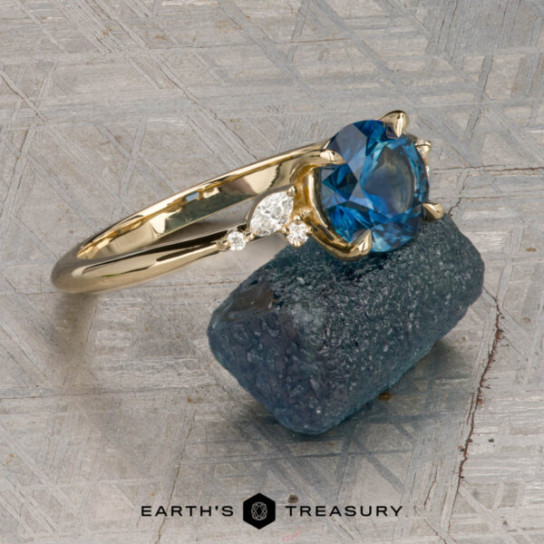The "Iris" ring in 14K yellow gold with 1.60-carat Montana sapphire The "Iris" ring in 14K yellow gold with a 1.60-carat Montana sapphire stands upright against a rough blue stone, with the "Earth's Treasury" logo below on a textured background.