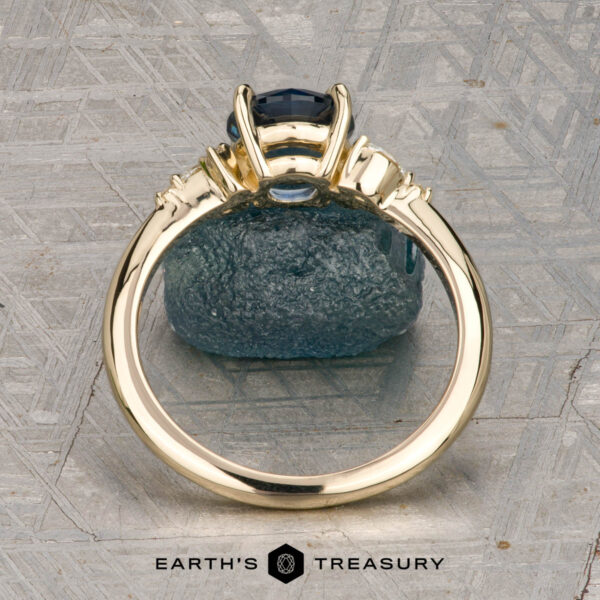 The "Iris" ring in 14K yellow gold with 1.60-carat Montana sapphire The "Iris" ring in 14K yellow gold with a 1.60-carat Montana sapphire stands upright against a rough blue stone, with the "Earth's Treasury" logo below on a textured background.