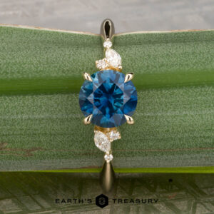 The "Iris" ring in 14K yellow gold, set with a 1.60-carat Montana sapphire and four white accent stones, rests on a green leaf; the Earth's Treasury logo is shown below.