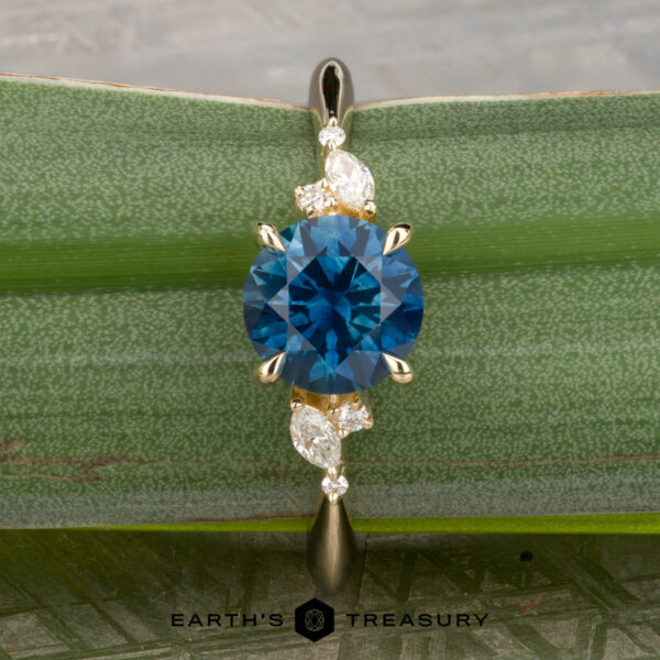 The "Iris" ring in 14K yellow gold with 1.60-carat Montana sapphire The "Iris" ring in 14K yellow gold, set with a 1.60-carat Montana sapphire and four white accent stones, rests on a green leaf; the Earth's Treasury logo is shown below.