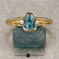 The "Madi" ring in 18K yellow gold with a 1.41-carat Montana sapphire is showcased on a rough blue-green stone, with the Earth's Treasury logo beneath on a patterned background.