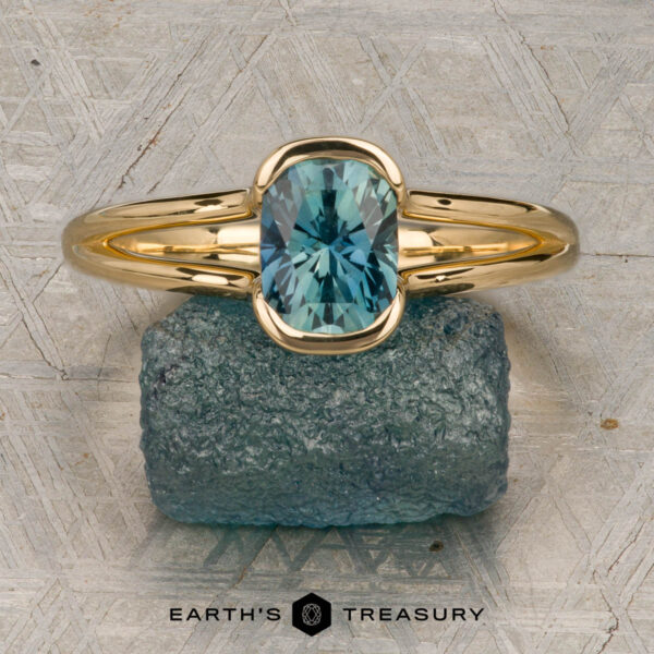 The "Madi" ring in 18K yellow gold with 1.41-carat Montana sapphire The "Madi" ring in 18K yellow gold with a 1.41-carat Montana sapphire is showcased on a rough blue-green stone, with the Earth's Treasury logo beneath on a patterned background.