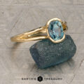 The "Madi" ring in 18K yellow gold with a 1.41-carat Montana sapphire is showcased on a rough blue-green stone, with the Earth's Treasury logo beneath on a patterned background.