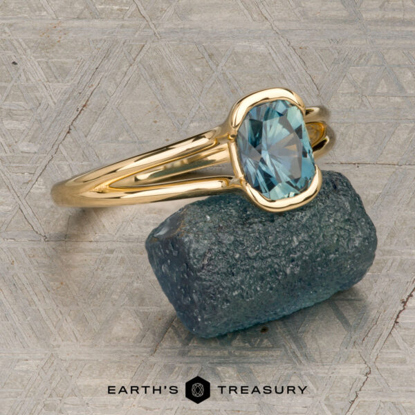 The "Madi" ring in 18K yellow gold with 1.41-carat Montana sapphire The "Madi" ring in 18K yellow gold with a 1.41-carat Montana sapphire is showcased on a rough blue-green stone, with the Earth's Treasury logo beneath on a patterned background.