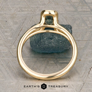 The "Madi" ring in 18K yellow gold with a 1.41-carat Montana sapphire is showcased on a rough blue-green stone, with the Earth's Treasury logo beneath on a patterned background.