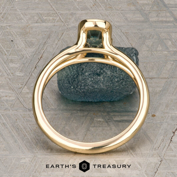 The "Madi" ring in 18K yellow gold with 1.41-carat Montana sapphire The "Madi" ring in 18K yellow gold with a 1.41-carat Montana sapphire is showcased on a rough blue-green stone, with the Earth's Treasury logo beneath on a patterned background.