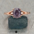 The "Marlowe" ring in 14K rose gold, featuring a 1.74-carat Umba sapphire, stands upright on a rough dark teal stone against a light patterned background.