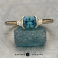 The "Dinara" ring in 14K yellow gold with a 1.14-carat Montana sapphire and two triangular clear stones rests on a rough blue mineral; Earth's Treasury logo appears below.