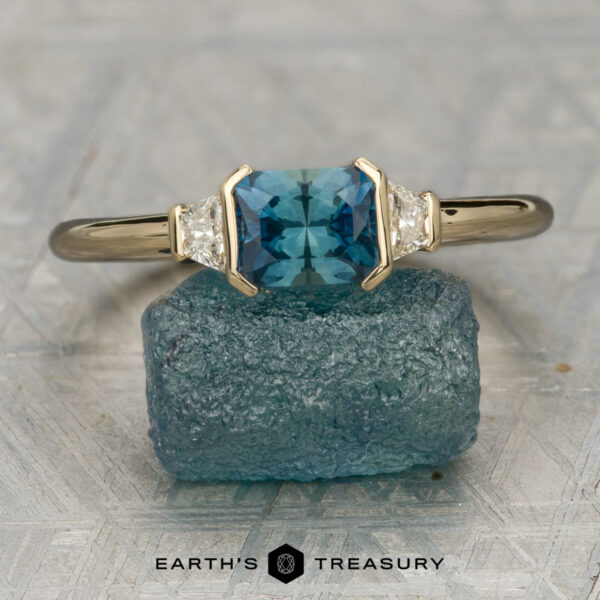The "Dinara" ring in 14K yellow gold with 1.14-carat Montana sapphire The "Dinara" ring in 14K yellow gold with a 1.14-carat Montana sapphire and two triangular clear stones rests on a rough blue mineral; Earth's Treasury logo appears below.