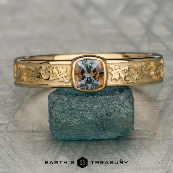 The "Kittley" ring in 18K yellow gold with 0.81-carat Montana sapphire The "Kittley" ring in 18K yellow gold features intricate floral engravings and a 0.81-carat Montana sapphire cushion-cut gemstone, elegantly displayed atop a textured greenish stone.
