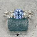 The "Yulania" ring showcases a platinum band with a 1.52-carat Ceylon sapphire center, accented by marquise white stones, displayed on a rough blue-green stone background.
