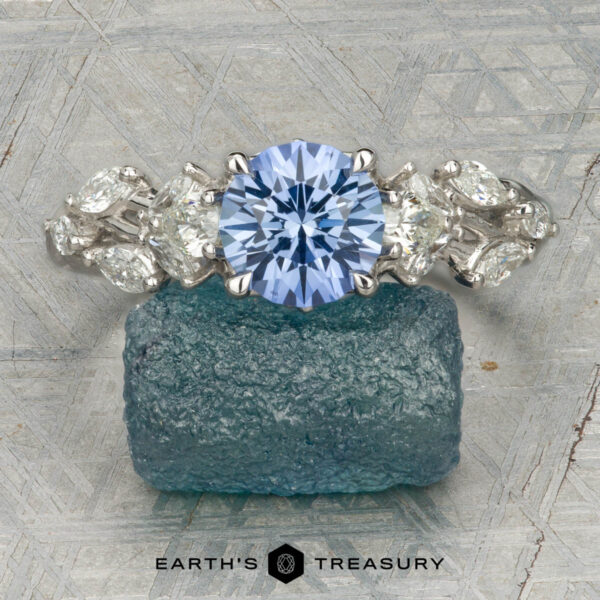 The "Yulania" ring in platinum with 1.52-carat Ceylon sapphire The "Yulania" ring showcases a platinum band with a 1.52-carat Ceylon sapphire center, accented by marquise white stones, displayed on a rough blue-green stone background.