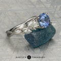 The "Yulania" ring showcases a platinum band with a 1.52-carat Ceylon sapphire center, accented by marquise white stones, displayed on a rough blue-green stone background.