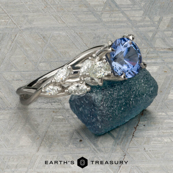 The "Yulania" ring in platinum with 1.52-carat Ceylon sapphire The "Yulania" ring showcases a platinum band with a 1.52-carat Ceylon sapphire center, accented by marquise white stones, displayed on a rough blue-green stone background.