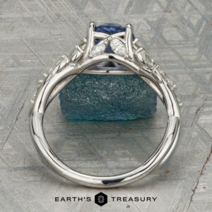 The "Yulania" ring showcases a platinum band with a 1.52-carat Ceylon sapphire center, accented by marquise white stones, displayed on a rough blue-green stone background.
