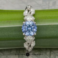 The "Yulania" ring features a platinum band with a 1.52-carat Ceylon sapphire at the center, accented by marquise and pear-shaped clear stones, beautifully displayed on a green leaf.