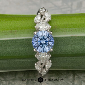 The "Yulania" ring features a platinum band with a 1.52-carat Ceylon sapphire at the center, accented by marquise and pear-shaped clear stones, beautifully displayed on a green leaf.