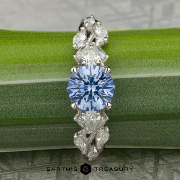 The "Yulania" ring features a platinum band with a 1.52-carat Ceylon sapphire at the center, accented by marquise and pear-shaped clear stones, beautifully displayed on a green leaf.