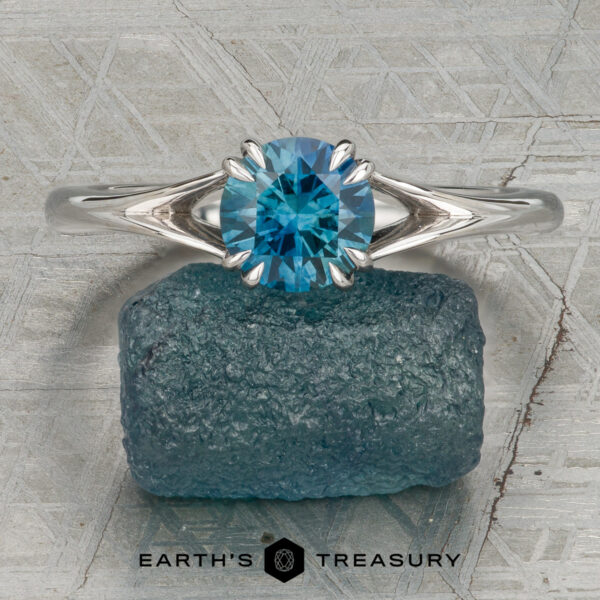 The Marlowe Ring The Marlowe ring with a round blue gemstone stands upright on a rough blue stone, set against a textured gray background. The text "Earth's Treasury" appears at the bottom.