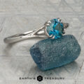 The Marlowe Ring featuring a blue gemstone, stacked from 53 images using Method=B (R=14, S=2), rests on a rough blue stone atop a textured surface with the Earth's Treasury logo below.