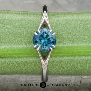 The Marlowe ring featuring a round blue gemstone in a platinum prong setting, shown on a green background with the "Earth's Treasury" logo at the bottom.