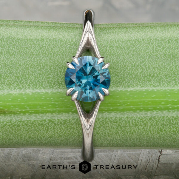 The Marlowe Ring The Marlowe ring featuring a round blue gemstone in a platinum prong setting, shown on a green background with the "Earth's Treasury" logo at the bottom.