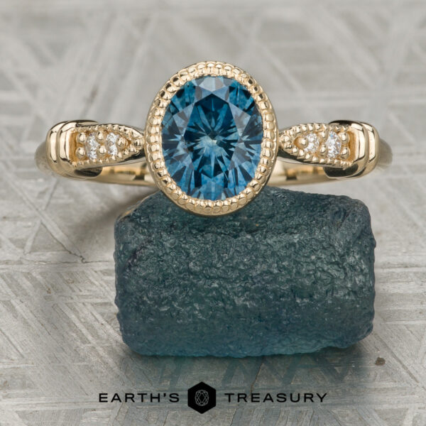 The “Myra” ring in 14K yellow gold with 1.57-carat Montana sapphire The “Myra” ring in 14K yellow gold features a 1.57-carat Montana sapphire and small diamonds, shown upright on a raw blue stone with a geometric-patterned background.