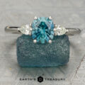 The Nymph Ring with two side diamonds is displayed on a rough blue gem, with "Earth's Treasury" and a logo visible at the bottom.