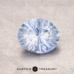 A 1.54-carat Ceylon Sapphire, round and light blue with a faceted surface, rests on a textured gray background with the text "Earth's Treasury" beneath it.