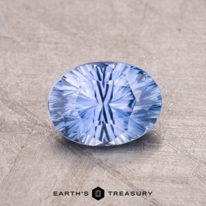 A 1.26-carat Ceylon Sapphire, a round, faceted blue gemstone, rests on a textured gray surface with the words "Earth's Treasury" beneath it.