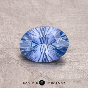 A 1.06-carat Ceylon Sapphire, round and faceted with intricate patterns on its textured blue surface, features Earth's Treasury's logo at the bottom.