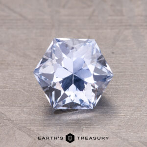 A 1.63-carat hexagonal Ceylon sapphire with a brilliant cut rests on a textured gray surface. Text below reads "Earth's Treasury.