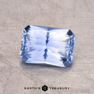 A 1.42-carat Ceylon sapphire, rectangular with faceted cuts, rests on a textured gray surface marked "Earth's Treasury.