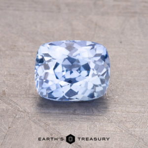 A 1.45-carat Ceylon Sapphire with a cushion cut, light blue hue rests on a textured gray surface.