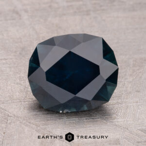 A 2.72-carat Montana Sapphire with a dark, deep blue hue and multiple facets rests on a textured surface. Below the gem, text reads "Earth's Treasury.