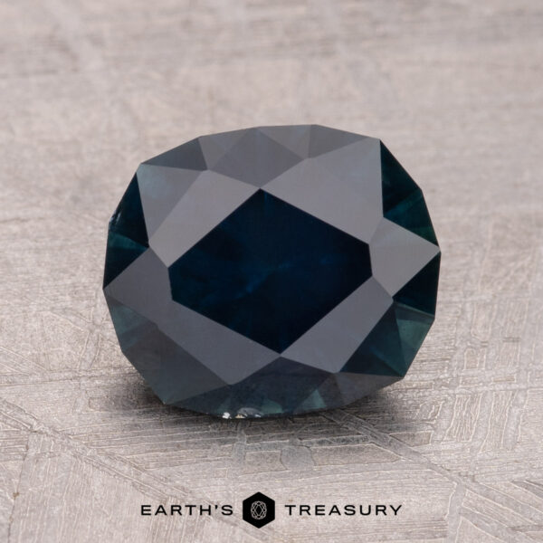 2.72-Carat Montana Sapphire (Heated) A 2.72-carat Montana Sapphire with a dark, deep blue hue and multiple facets rests on a textured surface. Below the gem, text reads "Earth's Treasury.